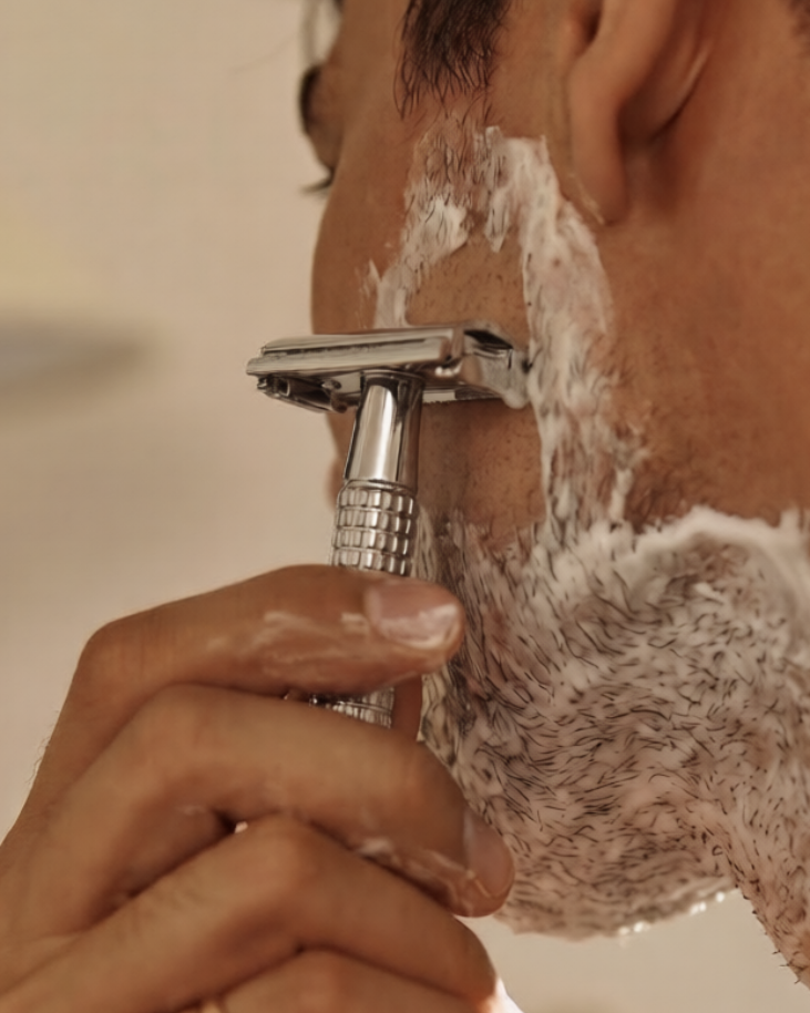Person shaving with a safety razor and shaving cream on a blurred background
