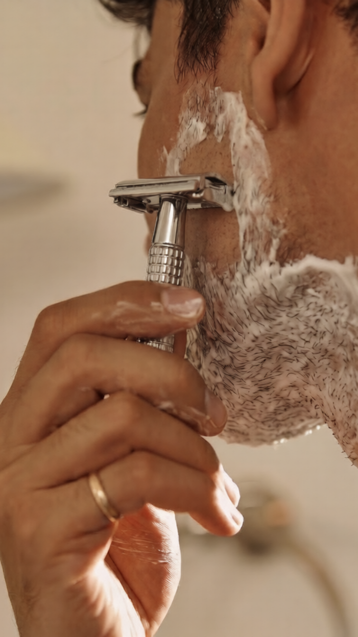 Person shaving with a safety razor and shaving cream on a blurred background