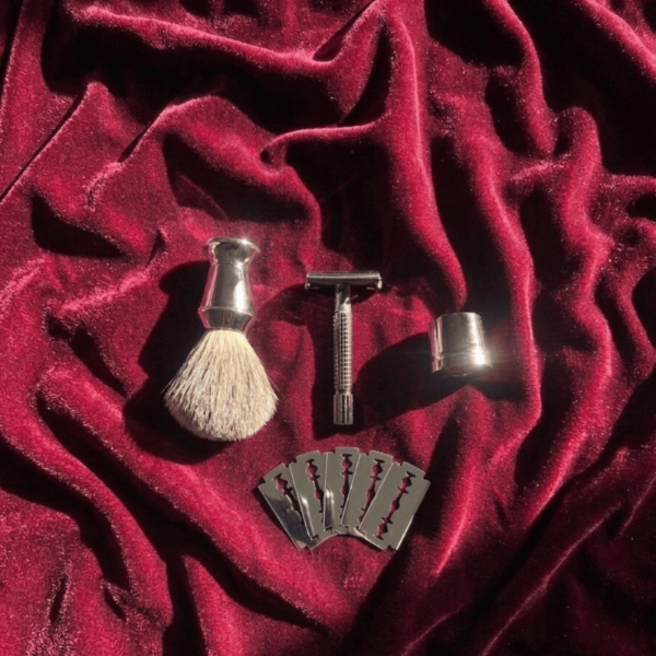 A vintage-style shaving set including a silver safety razor, shaving brush with cream bristles, metal cup, and four razor blades, arranged on a draped deep red velvet fabric.